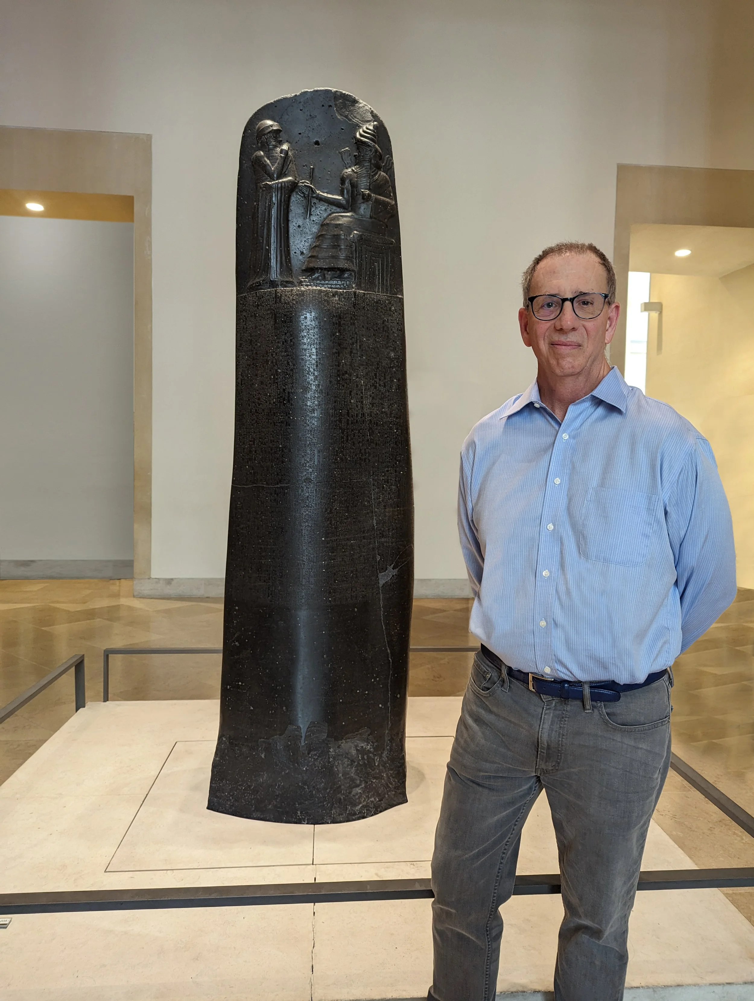 A man wearing glasses and a light blue shirt stands with his hands behind his back next to an ancient Egyptian black stone stele with hieroglyphic carvings displayed in a museum.