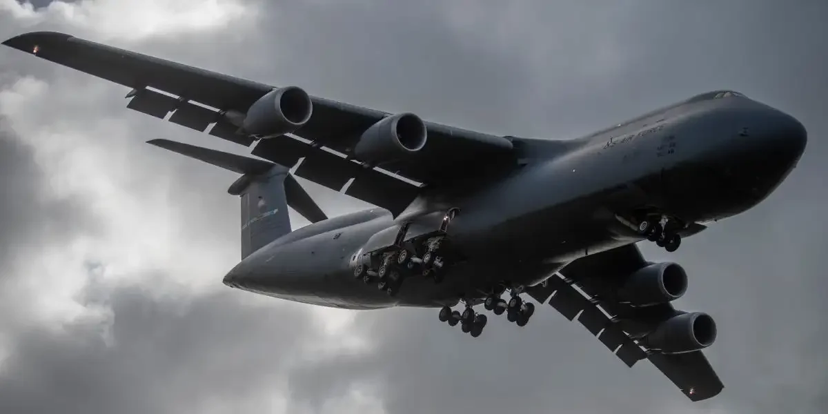A large military cargo plane flying under dark, cloudy sky.