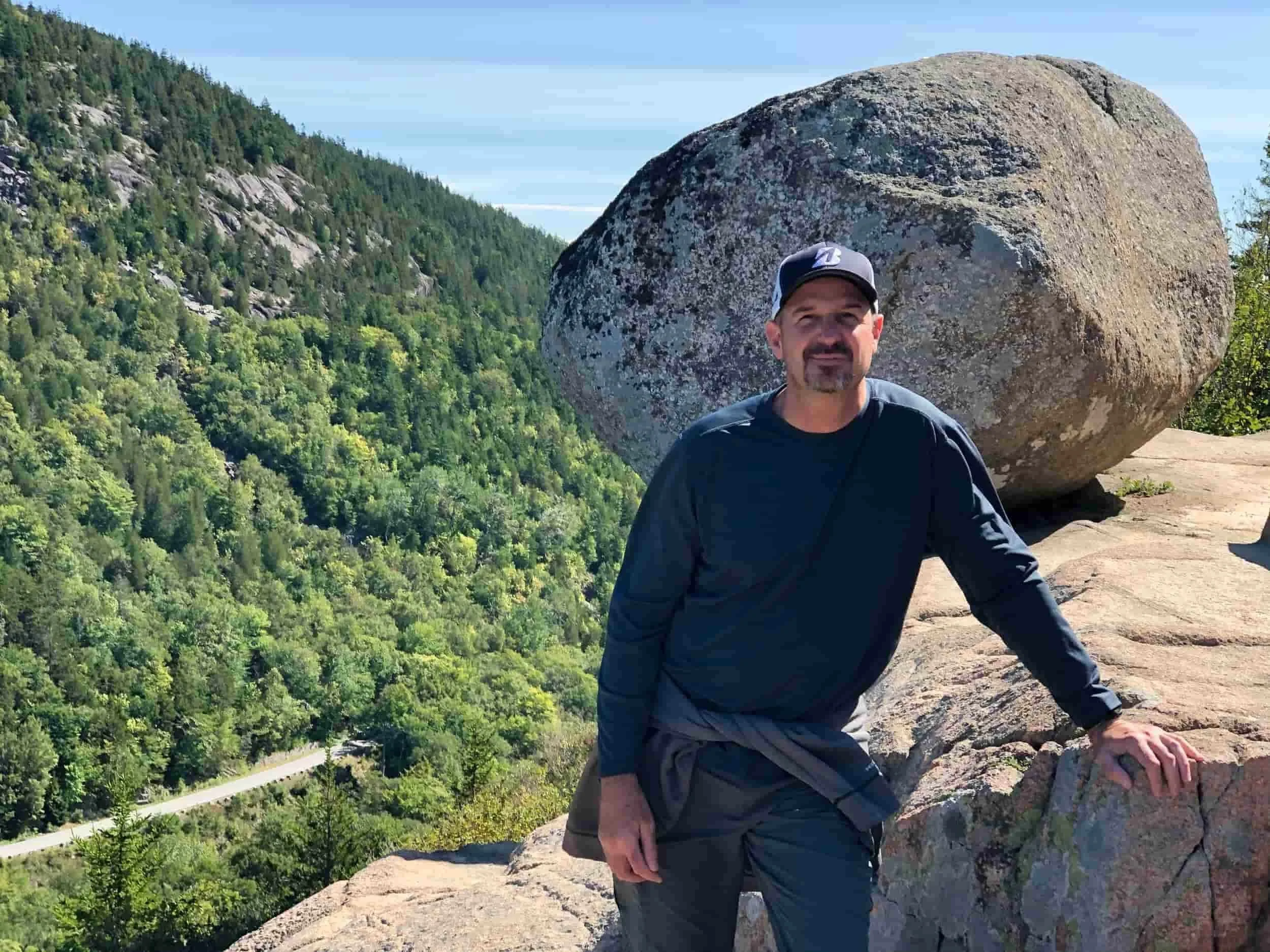 A man standing on a rocky ledge in front of a large boulder with a forested mountain in the background.