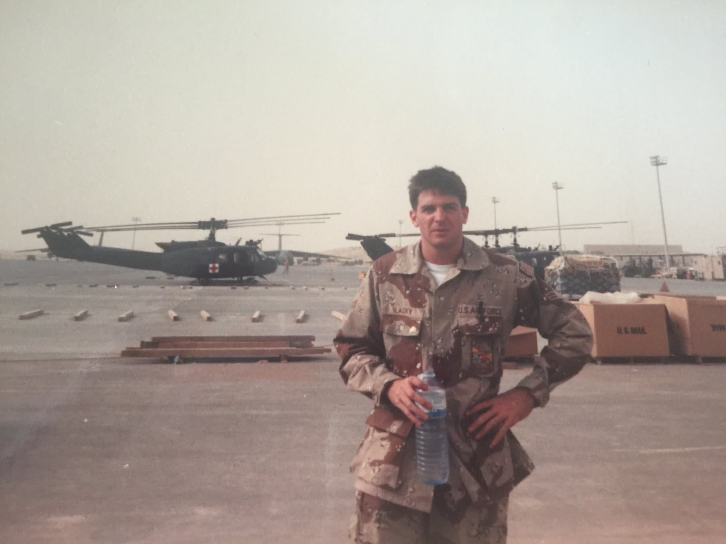 A young man in U.S. Army camouflage uniform standing in front of military helicopters on an airfield, holding a water bottle.