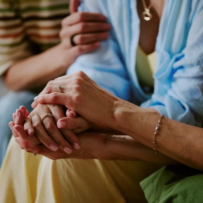 Close-up of two hands gently clasped, with one hand resting on top of the other, displaying rings and a bracelet, in a comforting gesture.
