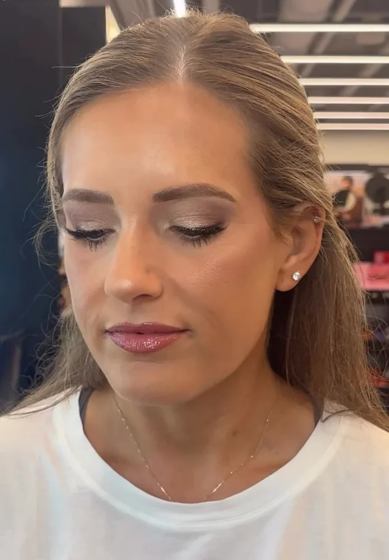 A woman with long light brown hair, looking down, wearing makeup, diamond earrings, a white top, and a delicate necklace, in a setting that appears to be a beauty or makeup studio.