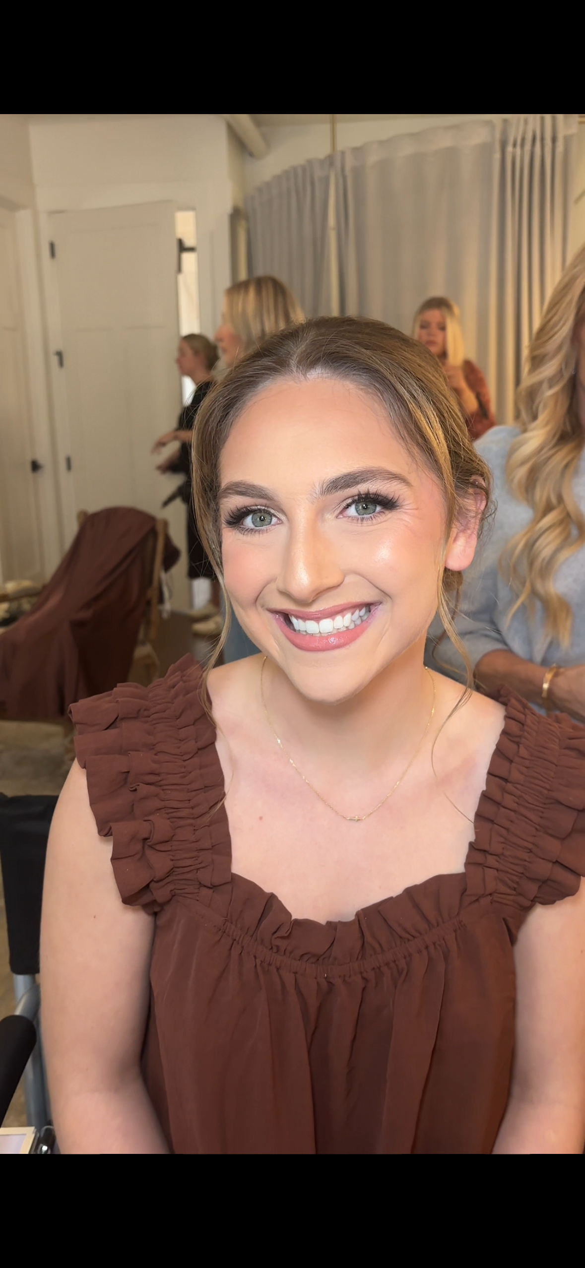 A young woman with light brown hair and blue eyes smiling at the camera, wearing a brown ruffled top and a delicate gold necklace. In the background, there are other women and a white curtain.