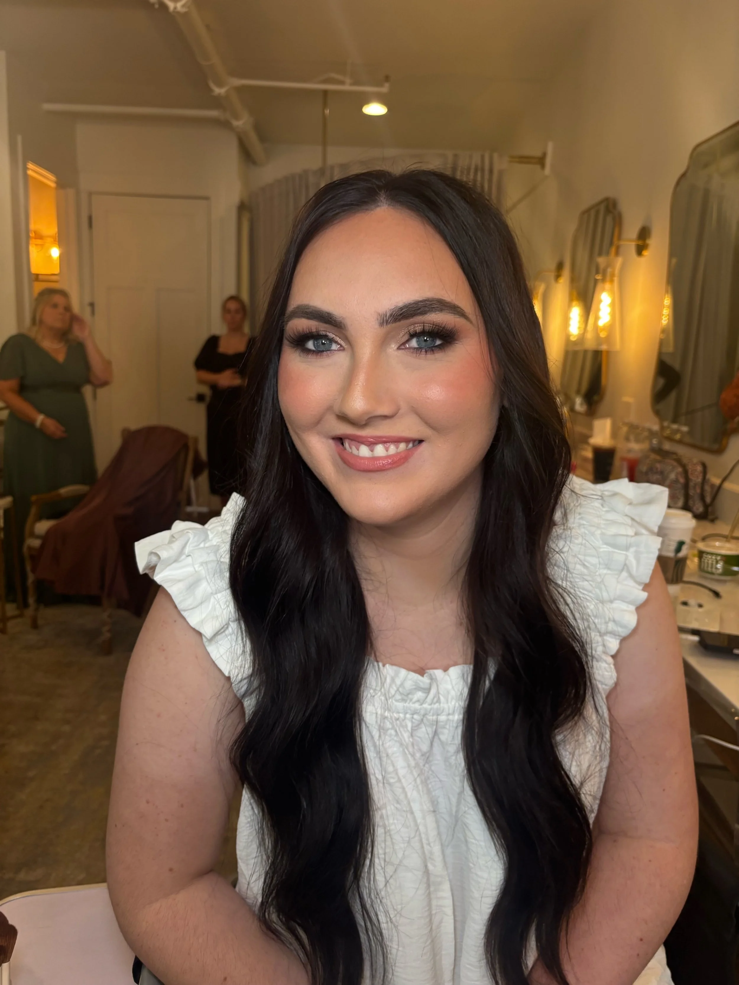 A young woman with long dark hair and blue eyes smiling at the camera, wearing a white ruffled top, in a warmly lit room with mirrors and other people in the background.