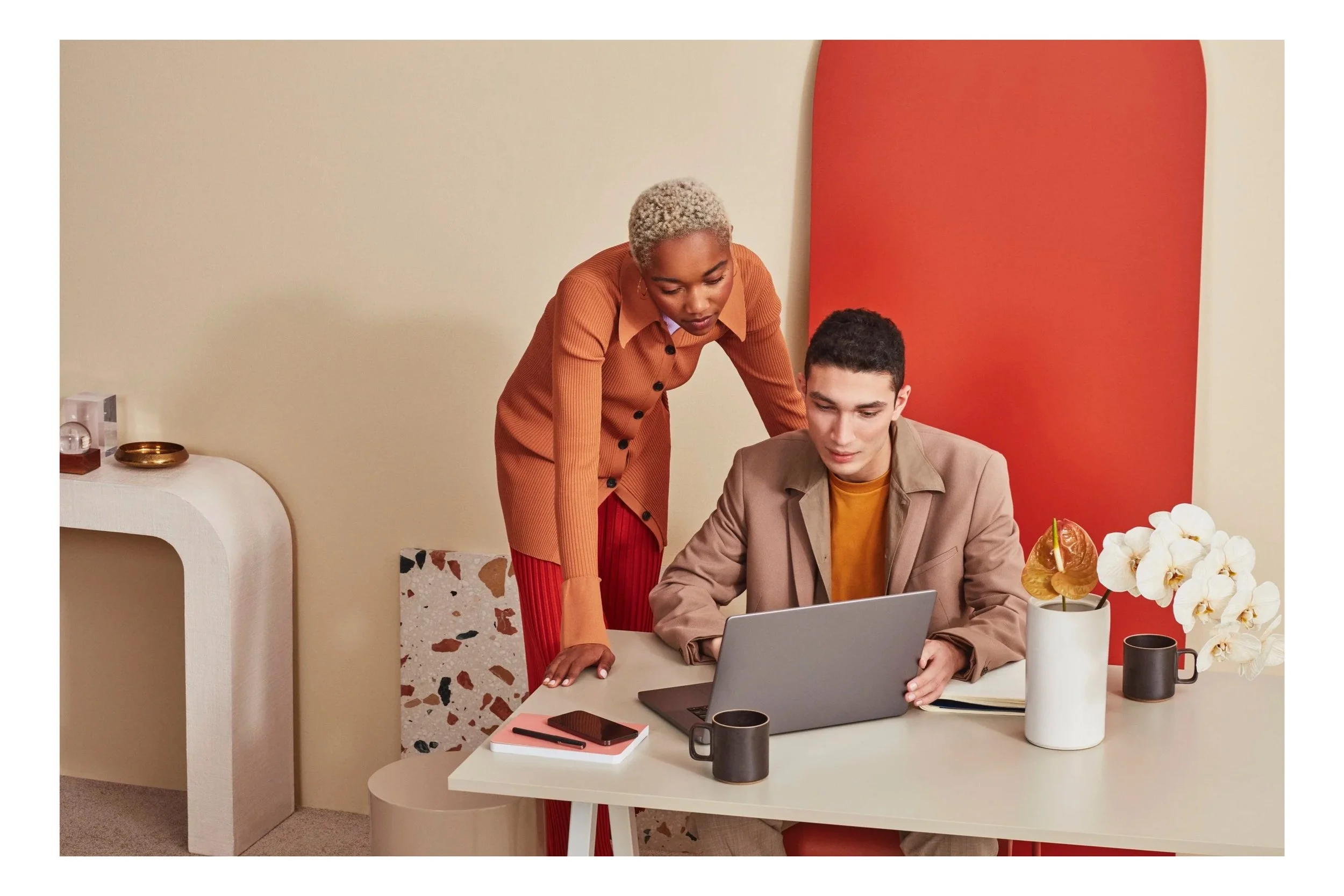 A woman with short, curly, bleach blonde hair leaning over a young man with dark hair sitting at a desk, looking at a laptop. The desk has a black mug, a notebook, a pen, and a potted orchid plant. The background features a red and beige wall.