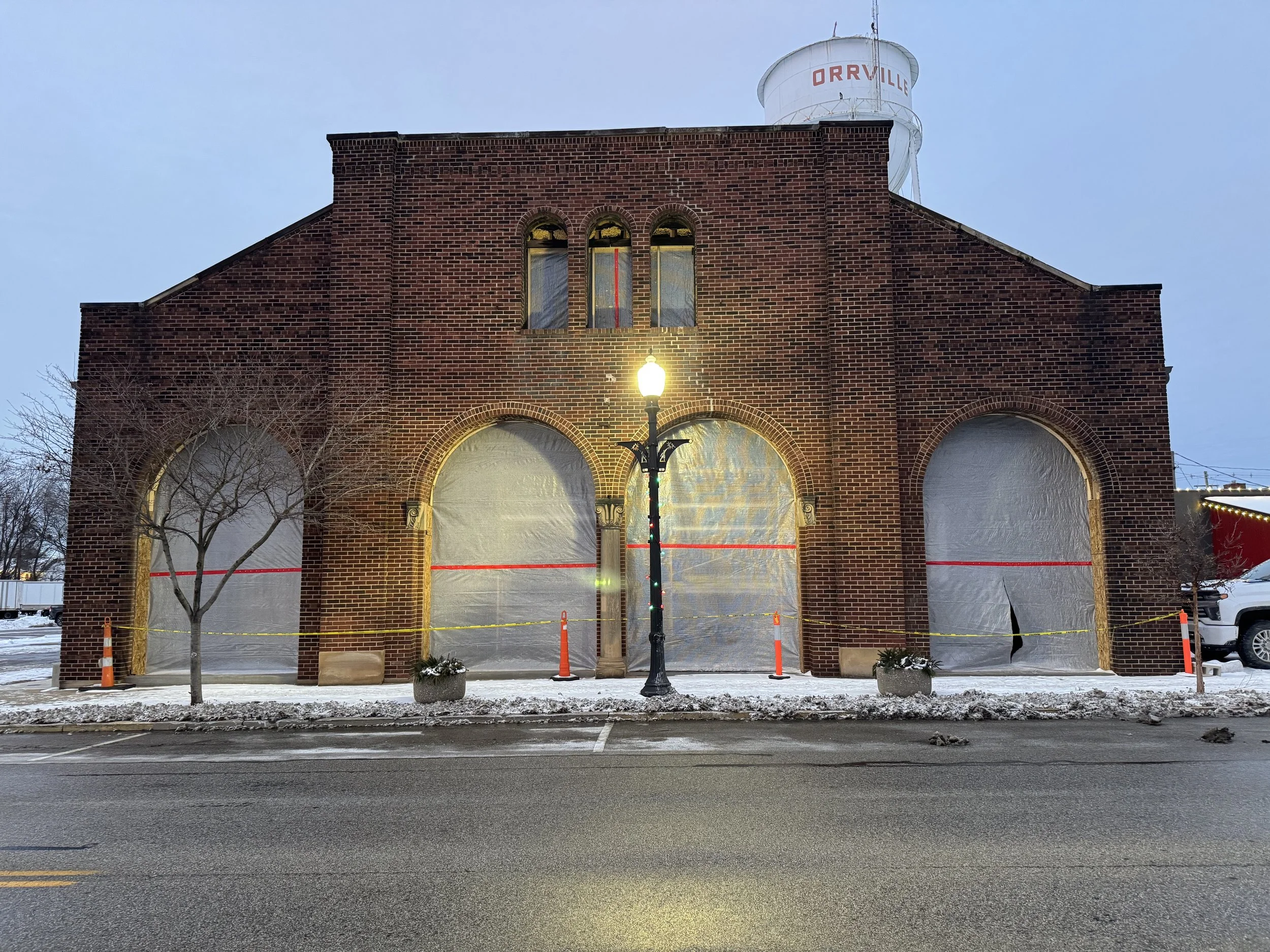 Old brick building with boarded-up windows decorated for Christmas, snow on the ground, a streetlamp in front, and a water tower on top with the word "ORRVILLE" on it.