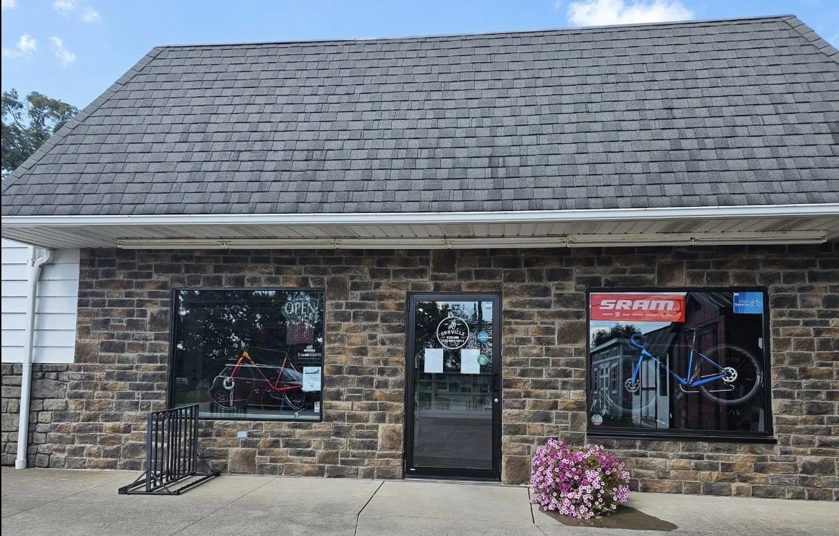 Front view of a brick bicycle shop with a stone facade, featuring two bike displays in windows, a glass door with a sign, and a potted flower outside.