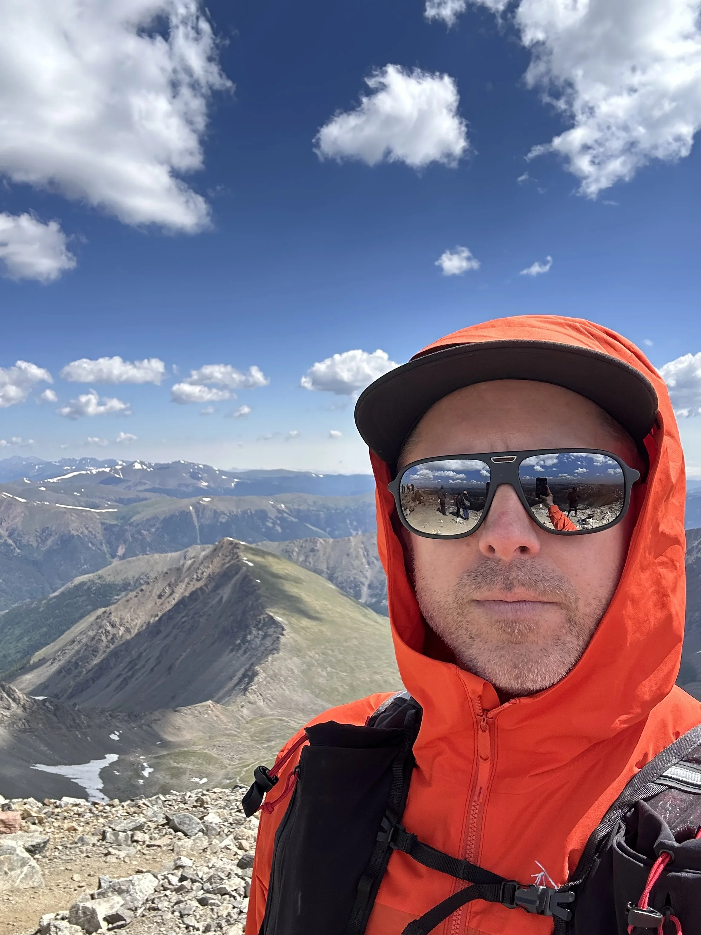 A man in orange outdoor gear and sunglasses taking a selfie on a mountain summit with a scenic view of rugged peaks, valleys, and snow patches under a partly cloudy sky.