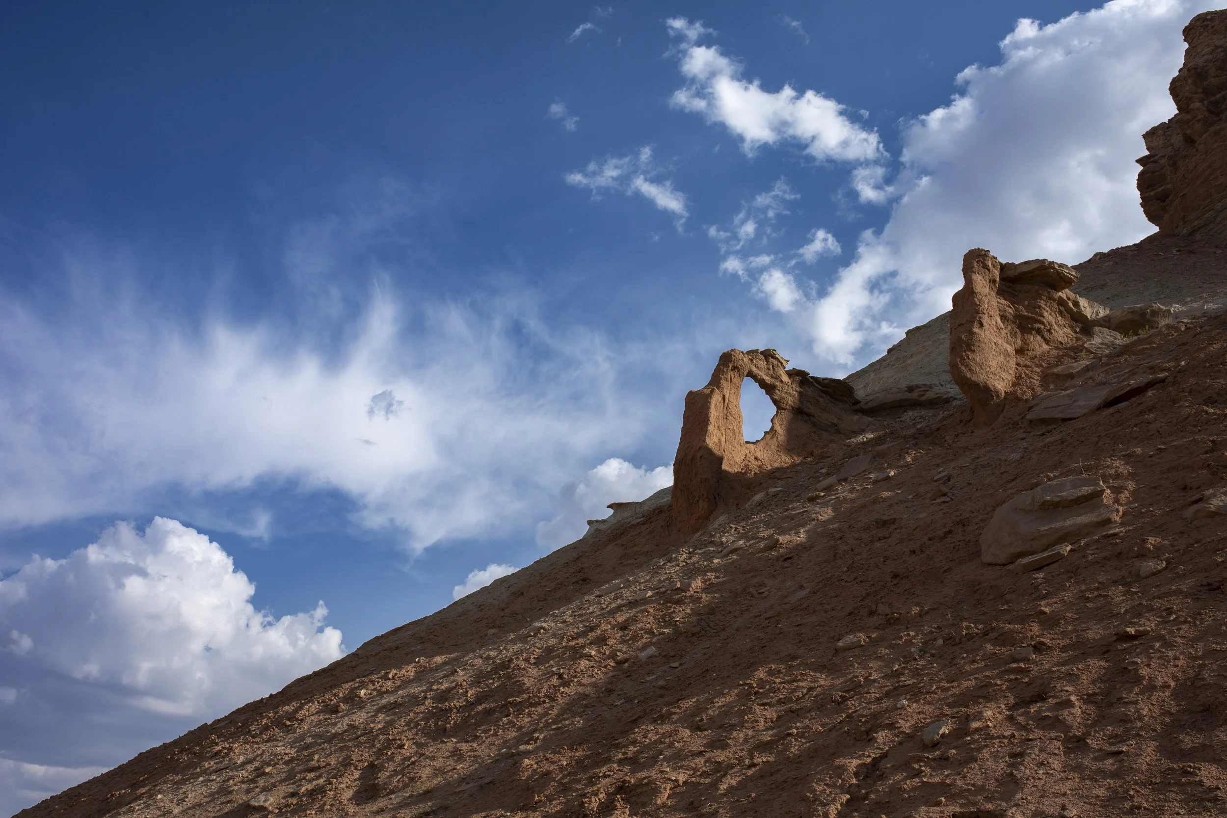 A natural rock formation resembling a bridge or arch on a sloped, desert landscape with a partly cloudy sky in the background.