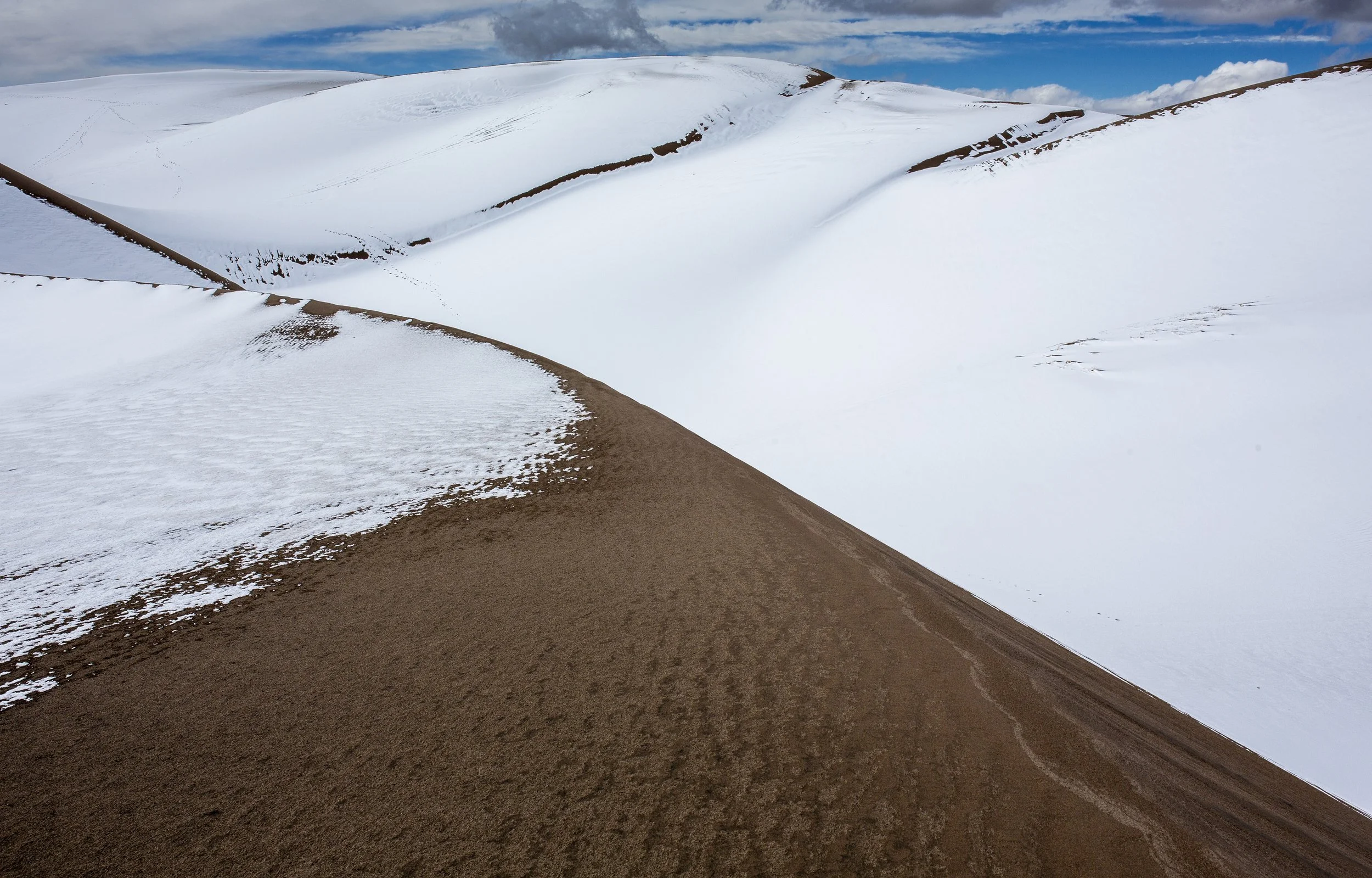 Snow-covered sand dune landscape with brown dirt slopes and swirling snow patterns under a partly cloudy sky.