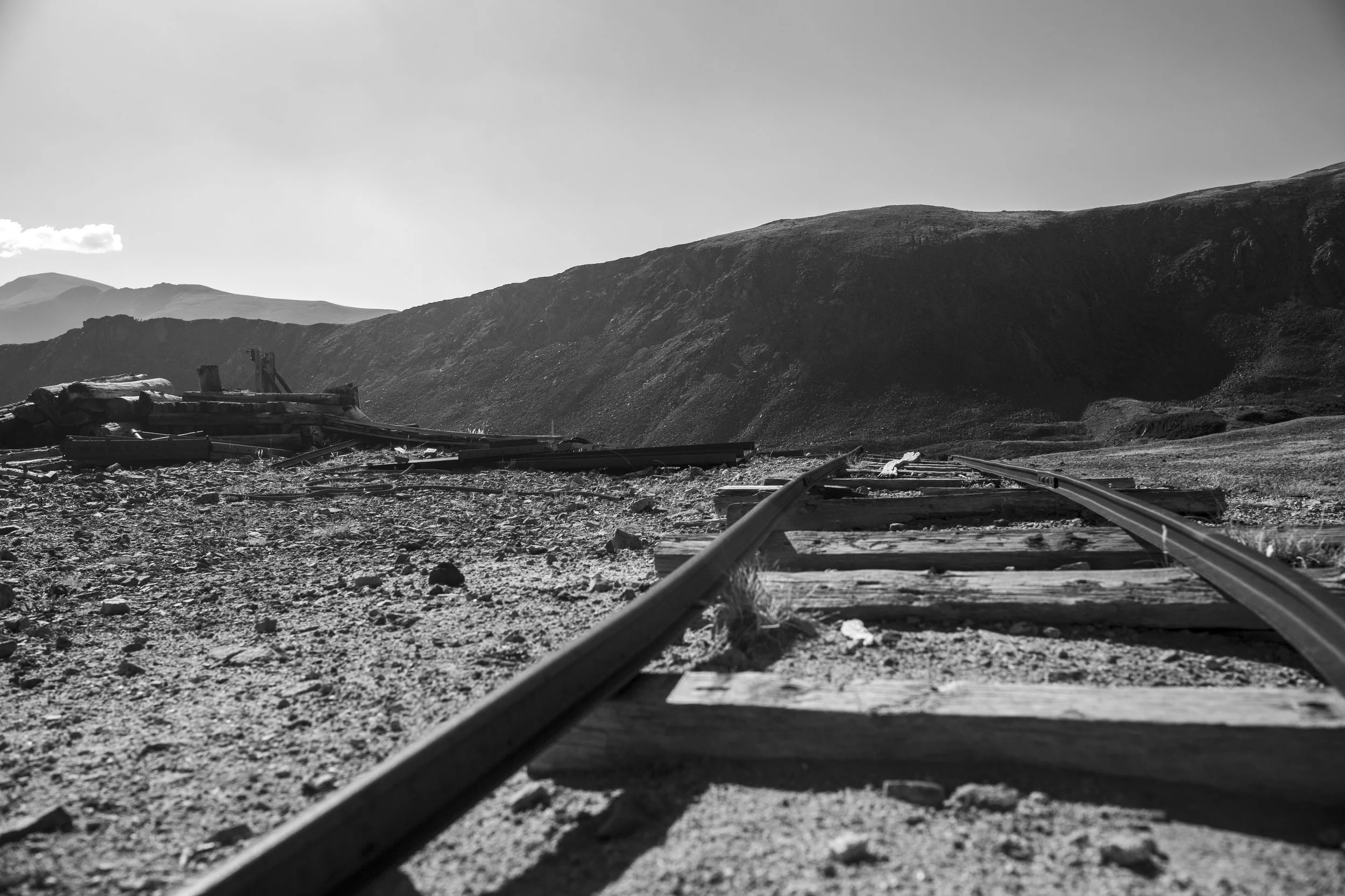 Black and white photograph of abandoned, broken railway tracks stretching into the distance in a barren landscape with hills or mountains in the background.