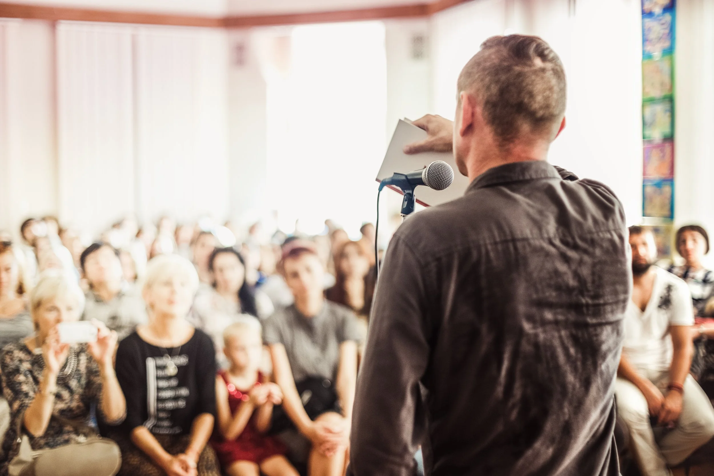 Man speaking into microphone in front of an audience during a presentation or event in a bright room.