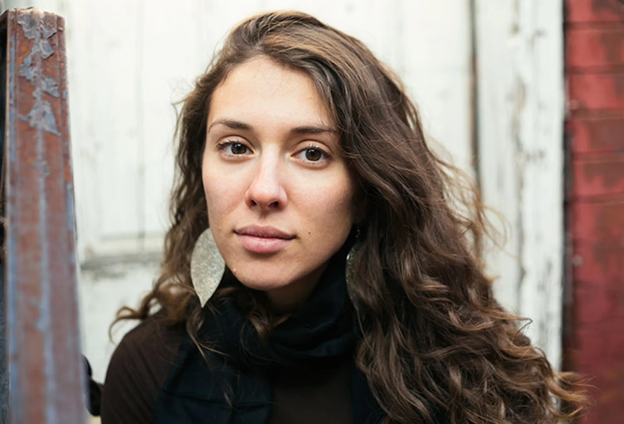 Close-up portrait of a young woman with long, wavy brown hair, wearing a black top and large silver earrings, standing outdoors near a weathered white and red wooden fence.