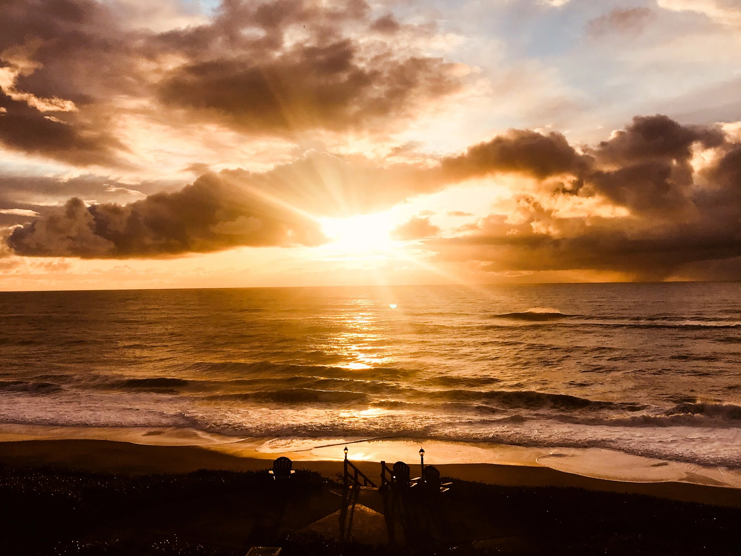 Sunset over the ocean with clouds in the sky, gentle waves on the shore, and three chairs on the beach facing the water.