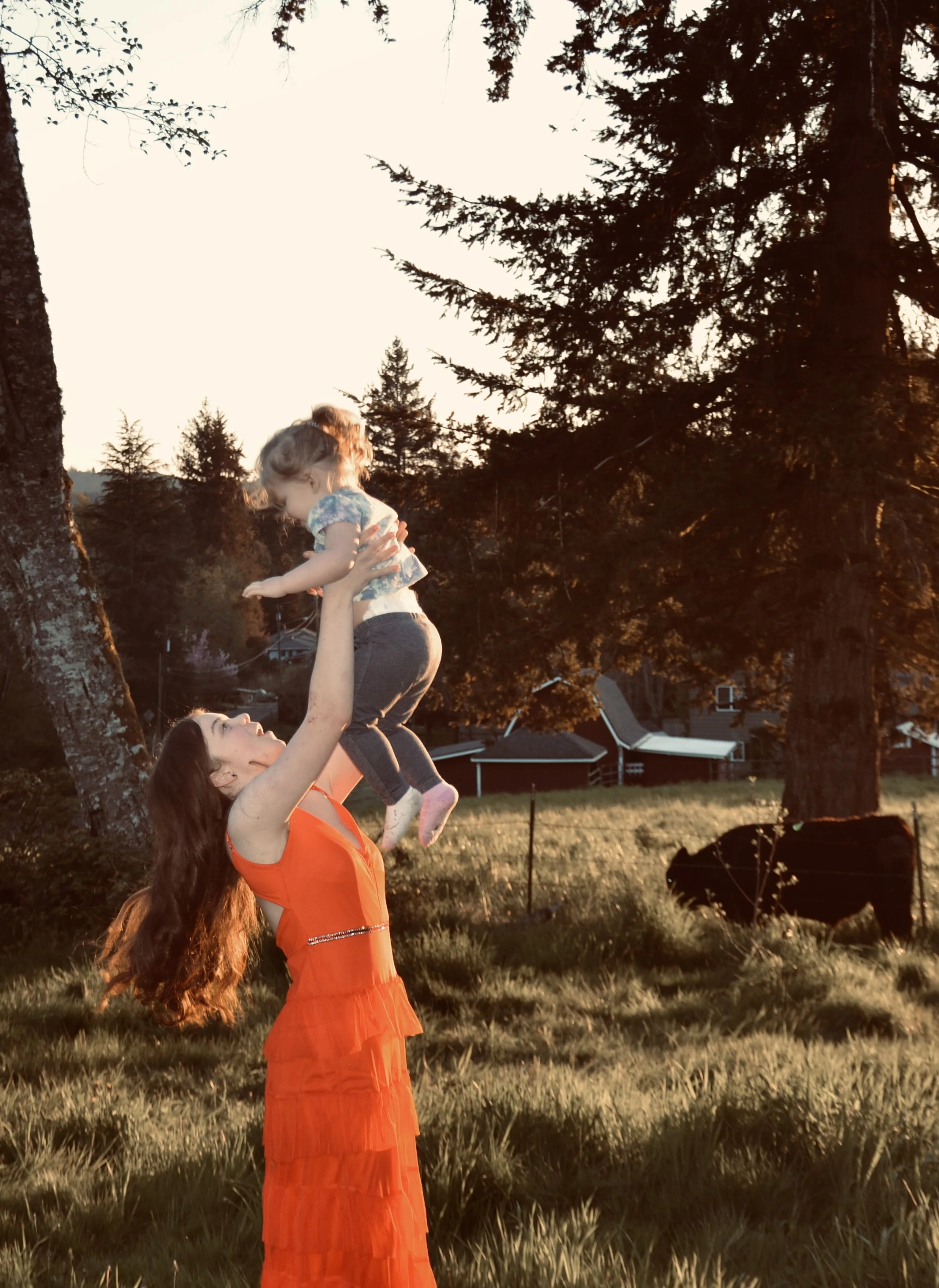 A woman with long reddish hair wearing an orange dress lifts a young girl with curly hair in a floral shirt and leggings into the air in a grassy field at sunset, with trees and a cow in the background.
