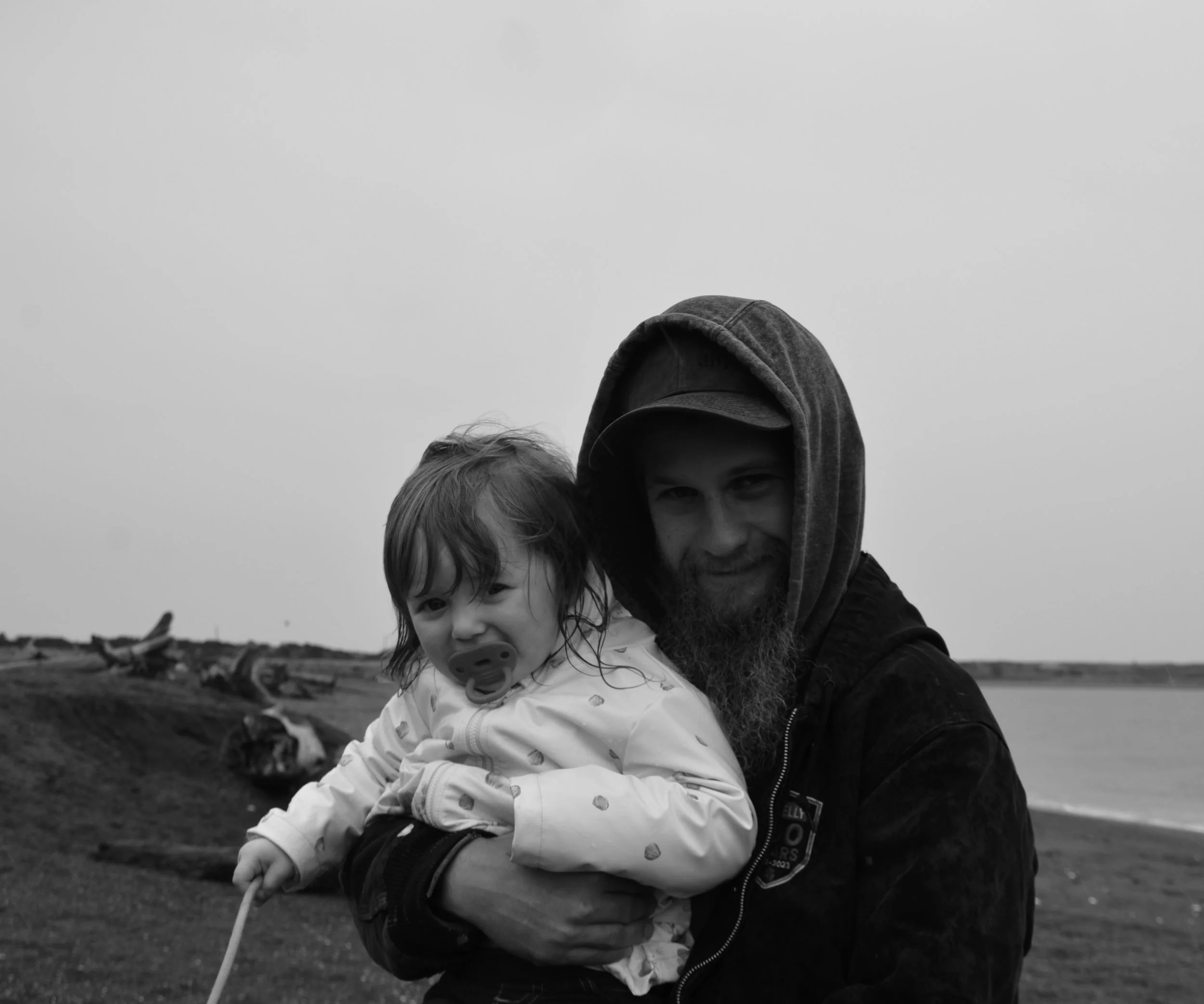 A man with a beard and hooded jacket holding a young girl with a pacifier, on a coastal landscape with driftwood and water in the background, in black and white.
