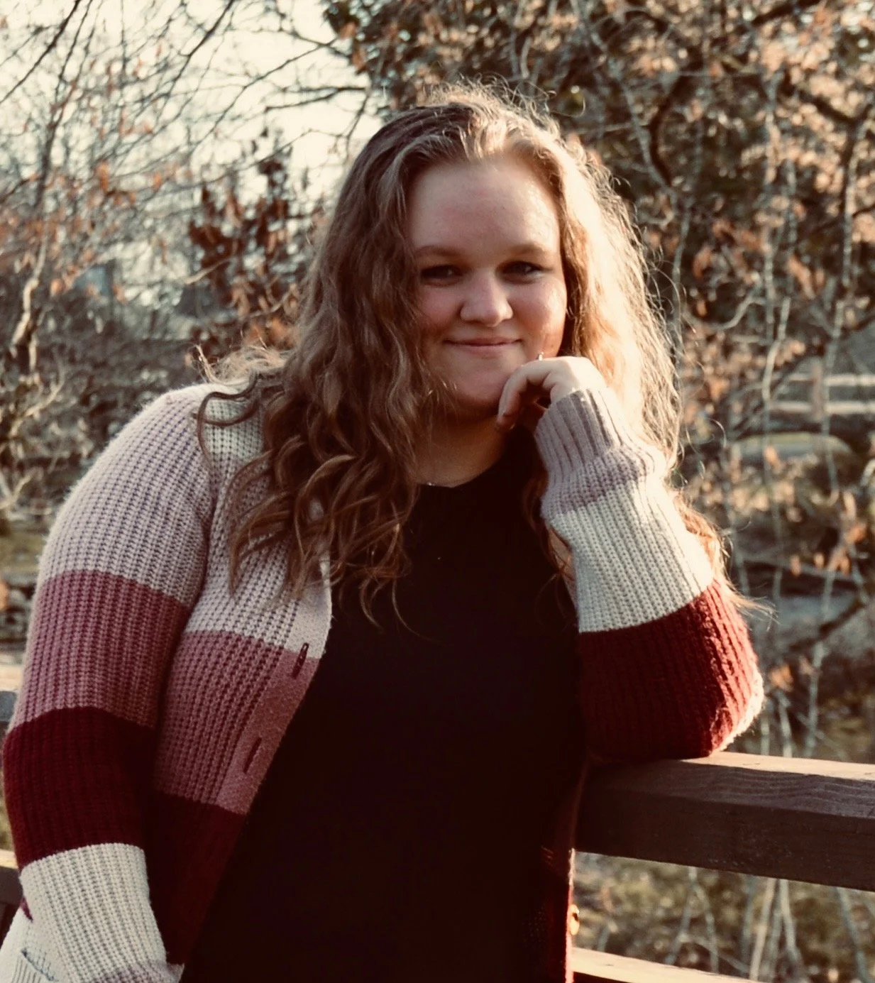 Young woman with curly hair leaning on a wooden railing outdoors during sunset, wearing a black top and a striped sweater, with trees in the background.