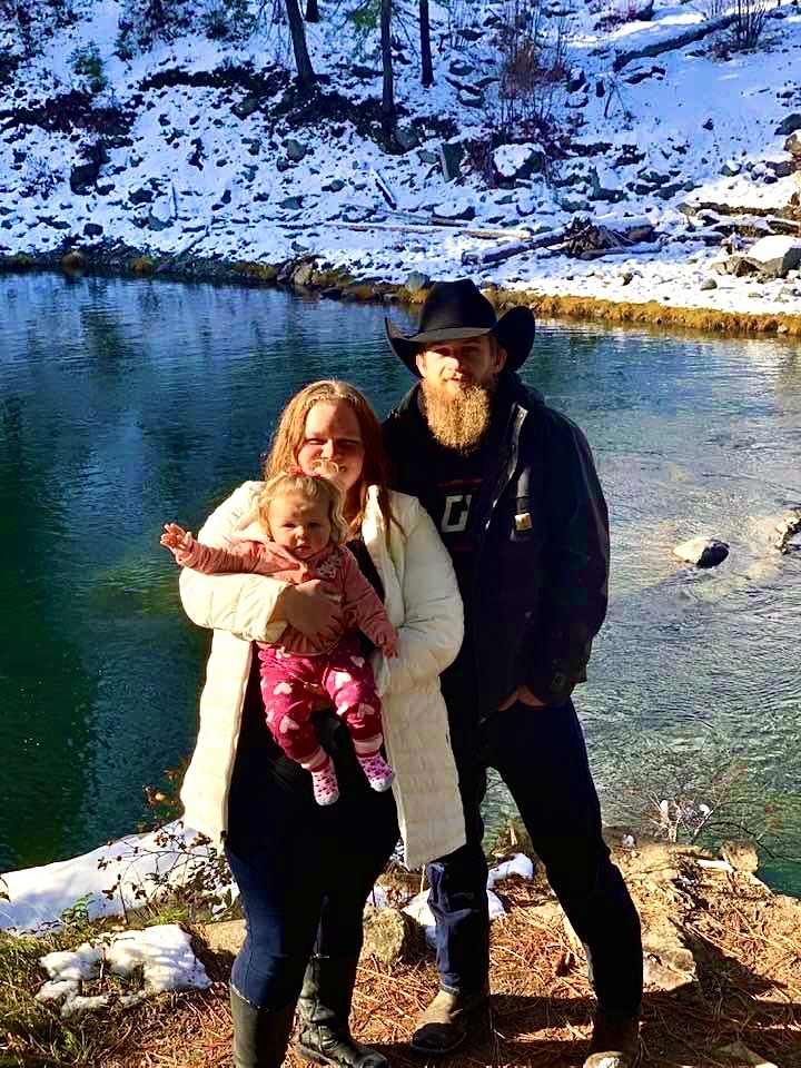 A family of three standing outdoors by a river with snow-covered ground in the background. The woman is holding a young girl, and the man wearing a cowboy hat stands beside them.