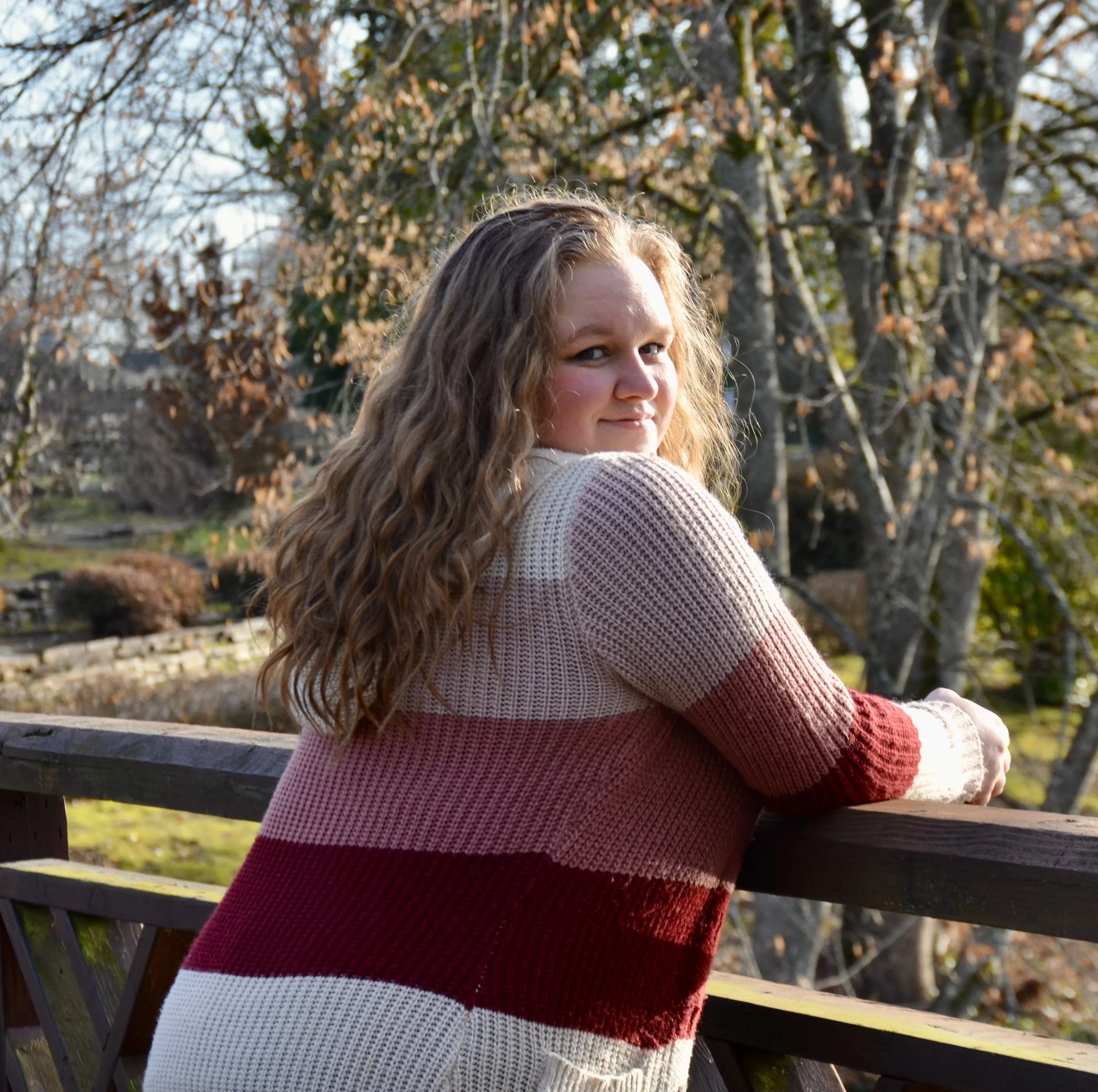 Woman with long curly hair leaning on a wooden railing outdoors during autumn.