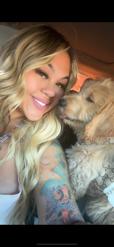 A woman with long blonde hair smiling next to a dog inside a car.