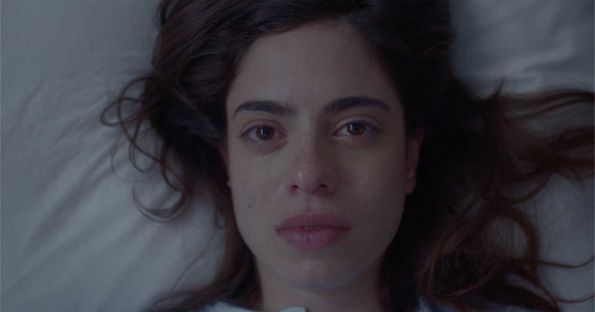 Close-up of a woman with light skin and brown hair lying on her back, looking directly at the camera, with a neutral expression.