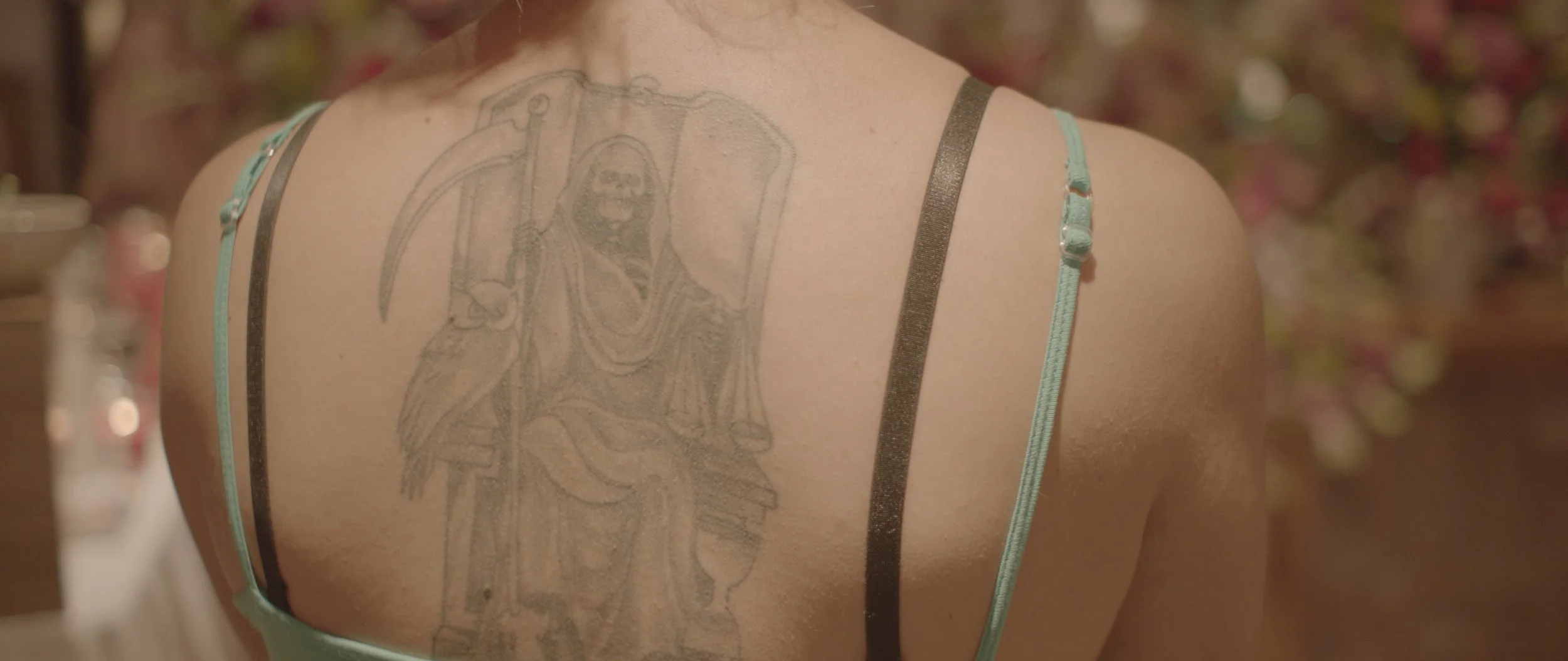 Back view of a person with a large tattoo of a Grim Reaper holding a scythe, wearing a hoodie, visible on their upper back and shoulder.