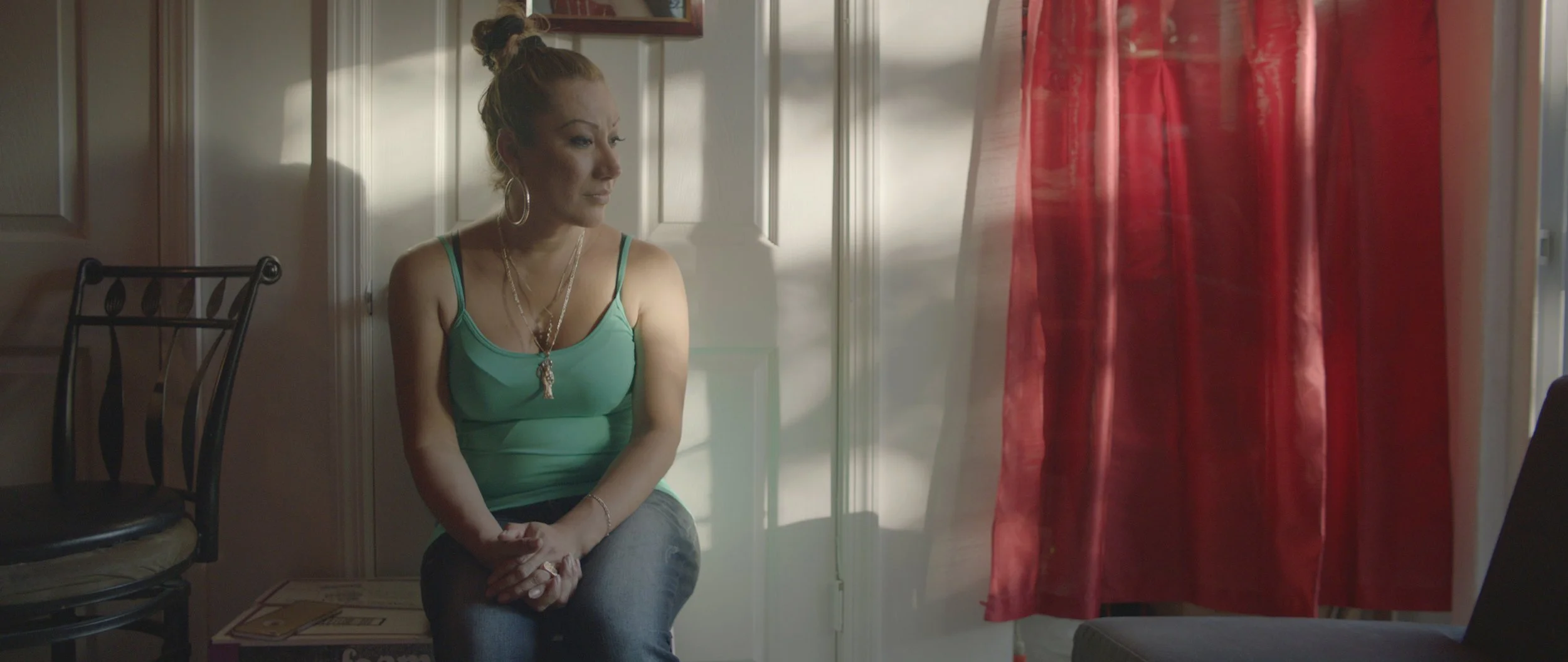 A woman with blonde hair tied in a bun, wearing a green tank top and jewelry, sitting by a window with red curtains, looking thoughtful.