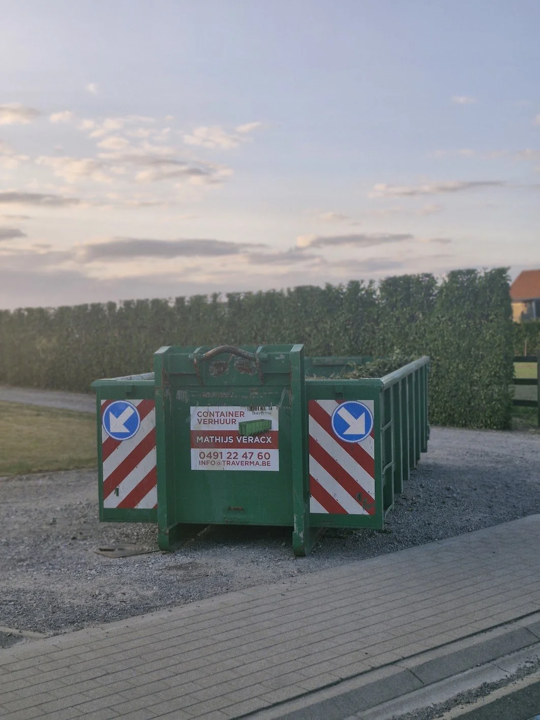 Green dumpster with a sign for container rental placed on a gravel surface next to a sidewalk with a rural background of trees and a house under a cloudy sky.