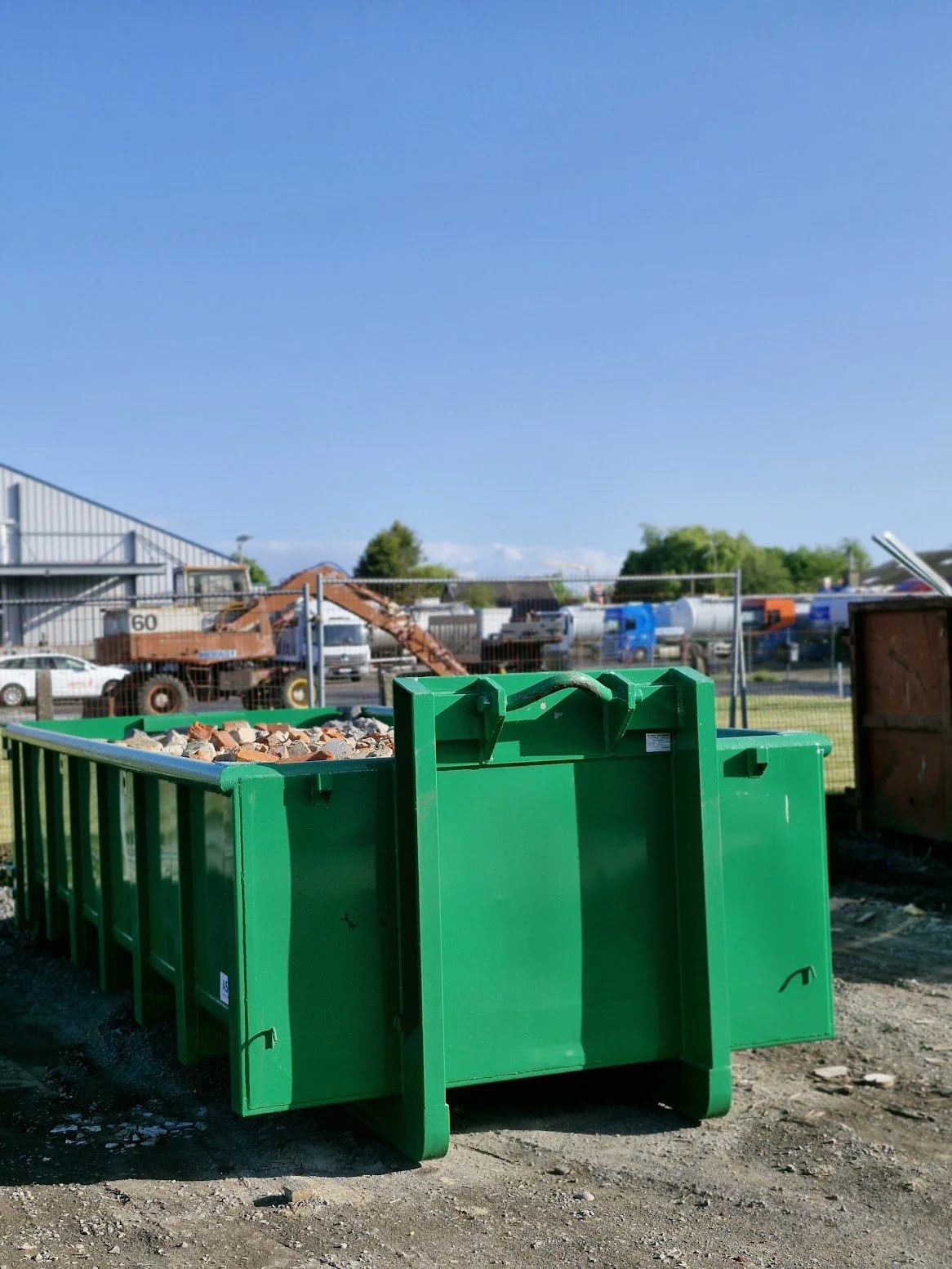 Green dumpster filled with rocks at a construction site with vehicles and industrial buildings in the background.