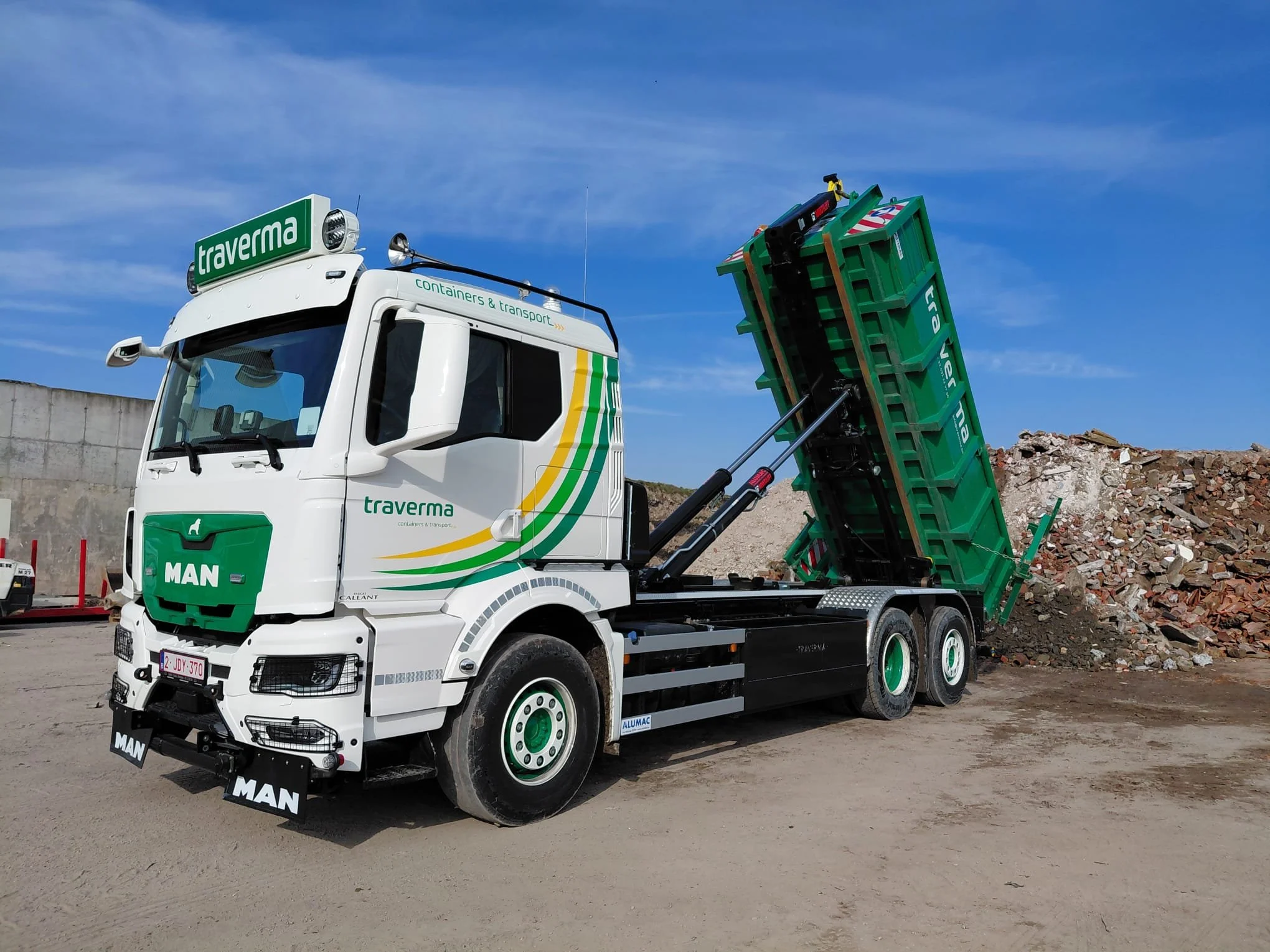 White truck with green and yellow accents lifting a large pile of rubble and rocks on a construction site, under a blue sky.