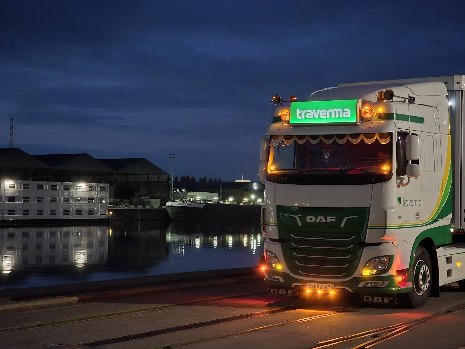 A large white and green DAF truck with a green illuminated 'traverma' sign on the front, parked near a waterfront at night, with industrial buildings, ships, and a cloudy sky in the background.