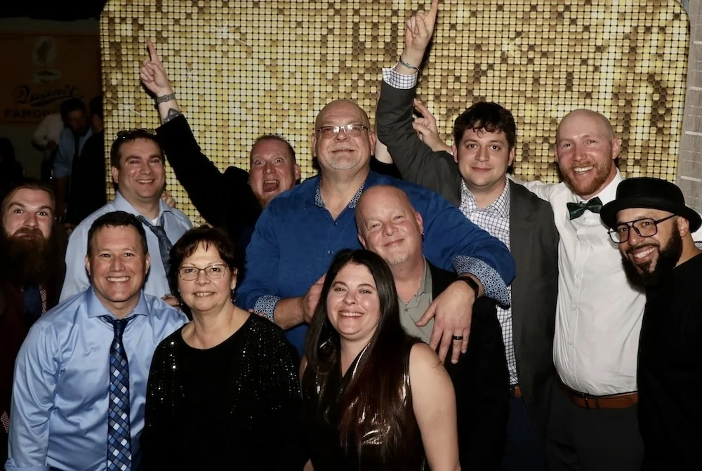 Group of ten people smiling and posing for a photo indoors, some making celebratory gestures, with a yellow mosaic wall in the background.