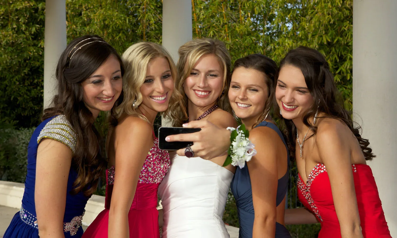 Group of six young women in colorful formal dresses taking a selfie together outdoors.