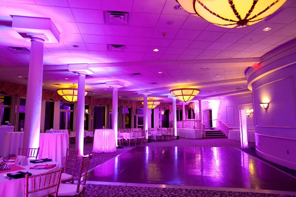 An elegant event venue with purple lighting, round chandeliers, white columns, and round tables with chairs, set up for a celebration or party.