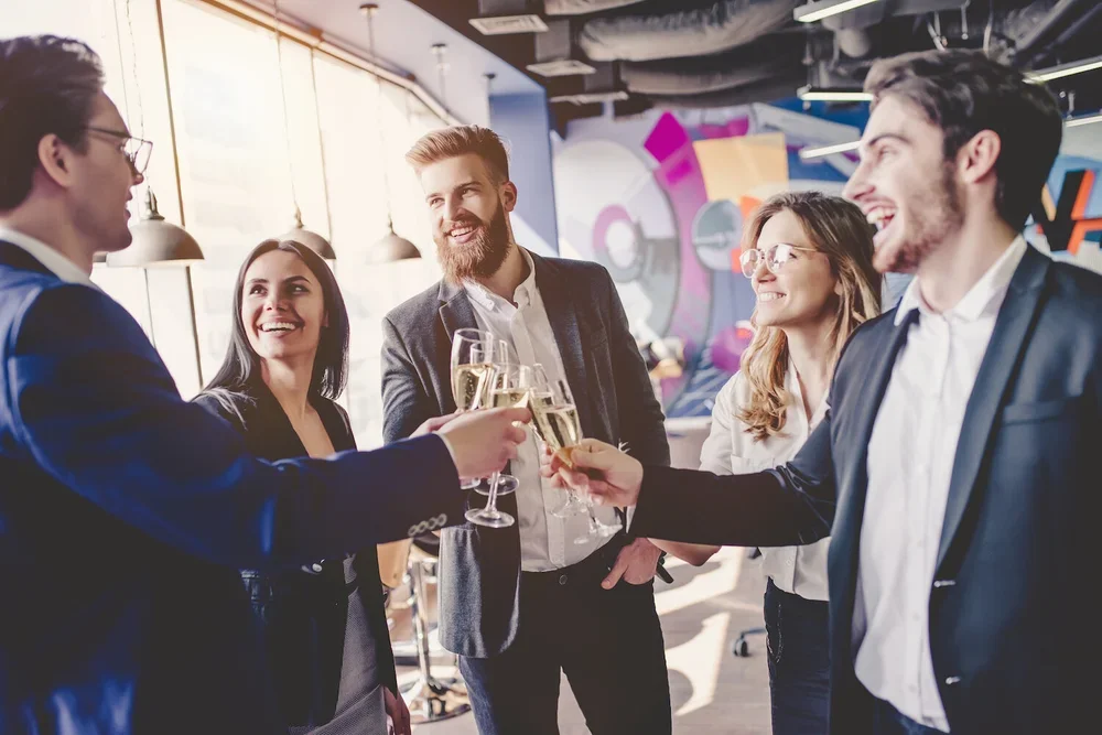 Group of five young professionals in business attire enjoying a toast with champagne in a modern office space.