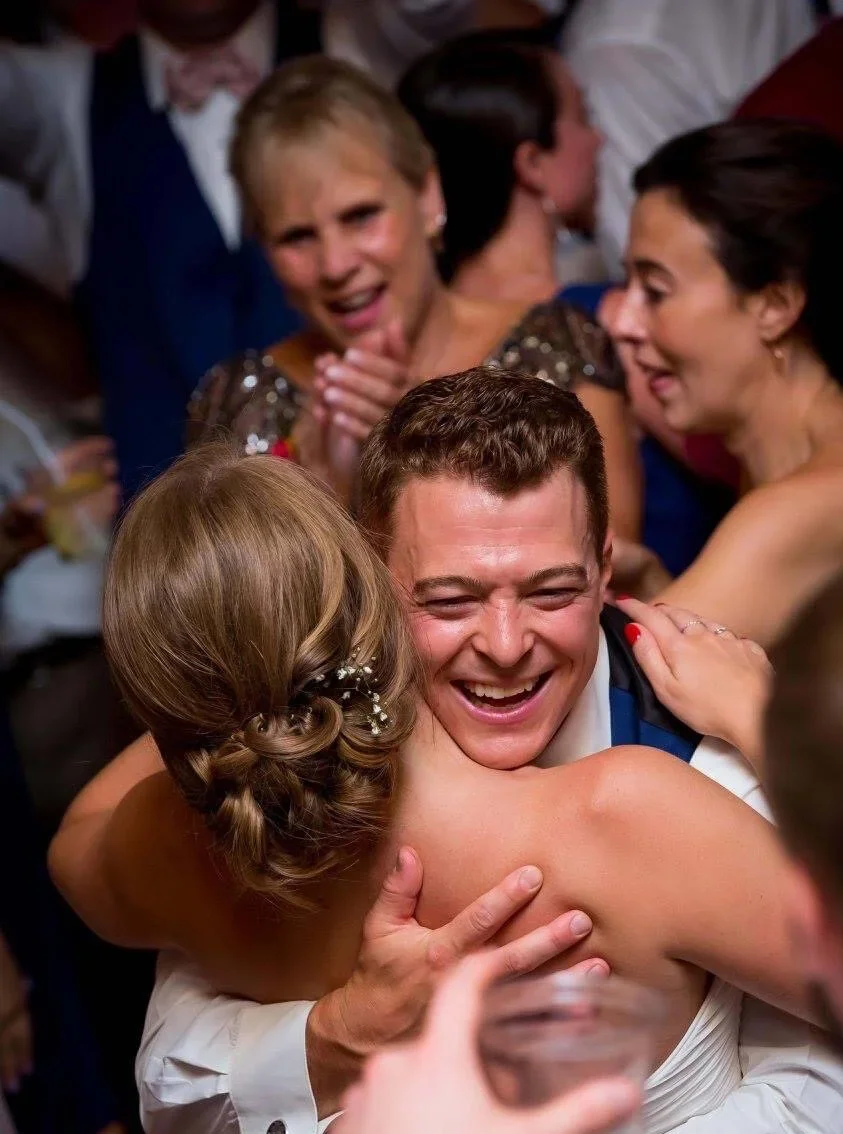 People at a celebration, with a man and woman hugging, smiling, and surrounded by others also enjoying the moment.
