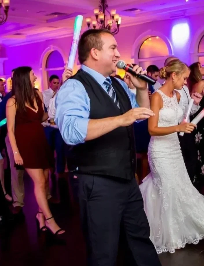A man in a suit and vest singing into a microphone at a wedding reception, with a bride in a white wedding gown holding a paper, and guests dancing in colorful lighting in the background.