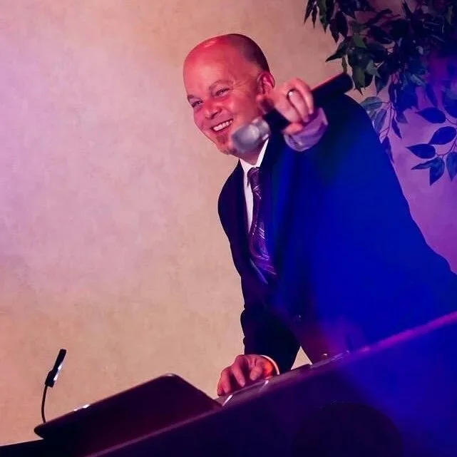 A man in a suit smiling and leaning over a podium, holding a microphone, with a plant and a plain wall in the background.
