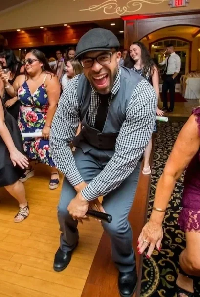 A man wearing glasses, a flat cap, and a checkered shirt smiling and holding a microphone while dancing at a social gathering with other smiling people in the background.