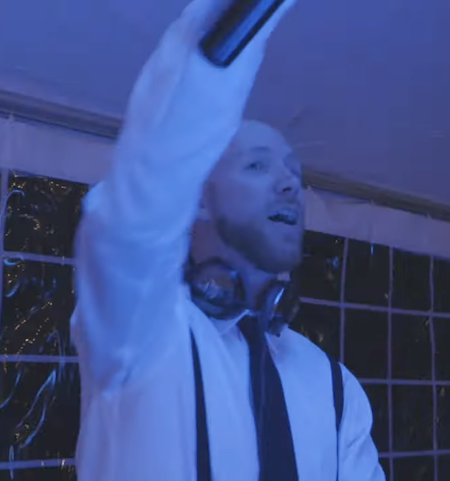 A DJ wearing a white shirt and headphones around his neck performs at a nightclub with blue lighting.