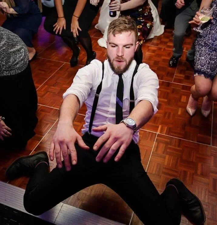 Man with a beard in a white shirt, black suspenders, and a black tie dancing on a wooden floor at a party, surrounded by other people holding drinks.