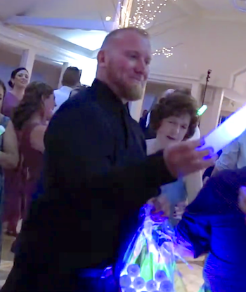 A smiling man in a dark suit holding a glowing stick at a party with other people and festive lighting.