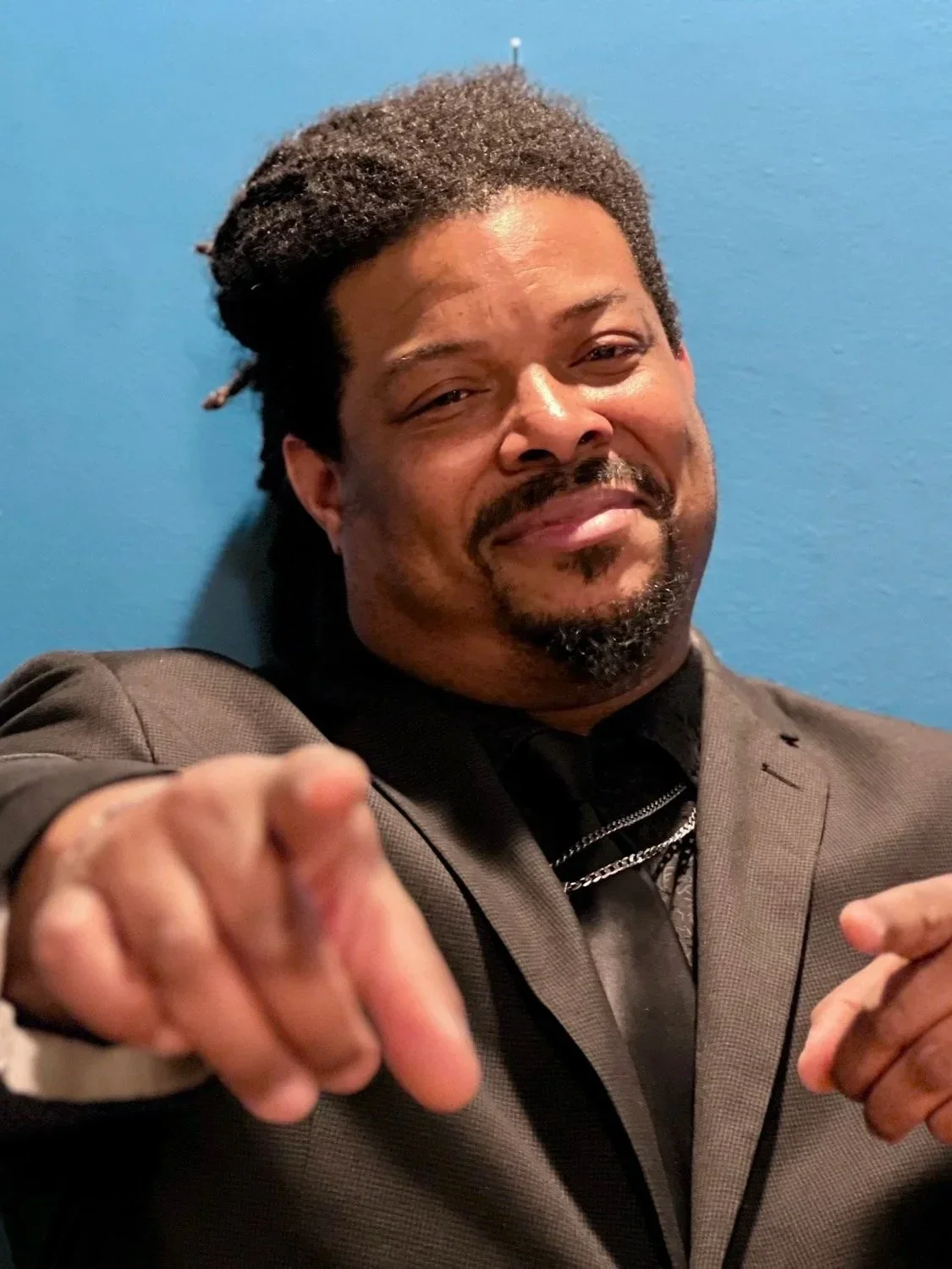 A man with a mustache and beard wearing a brown suit and black shirt pointing towards the camera, with a blue wall in the background.