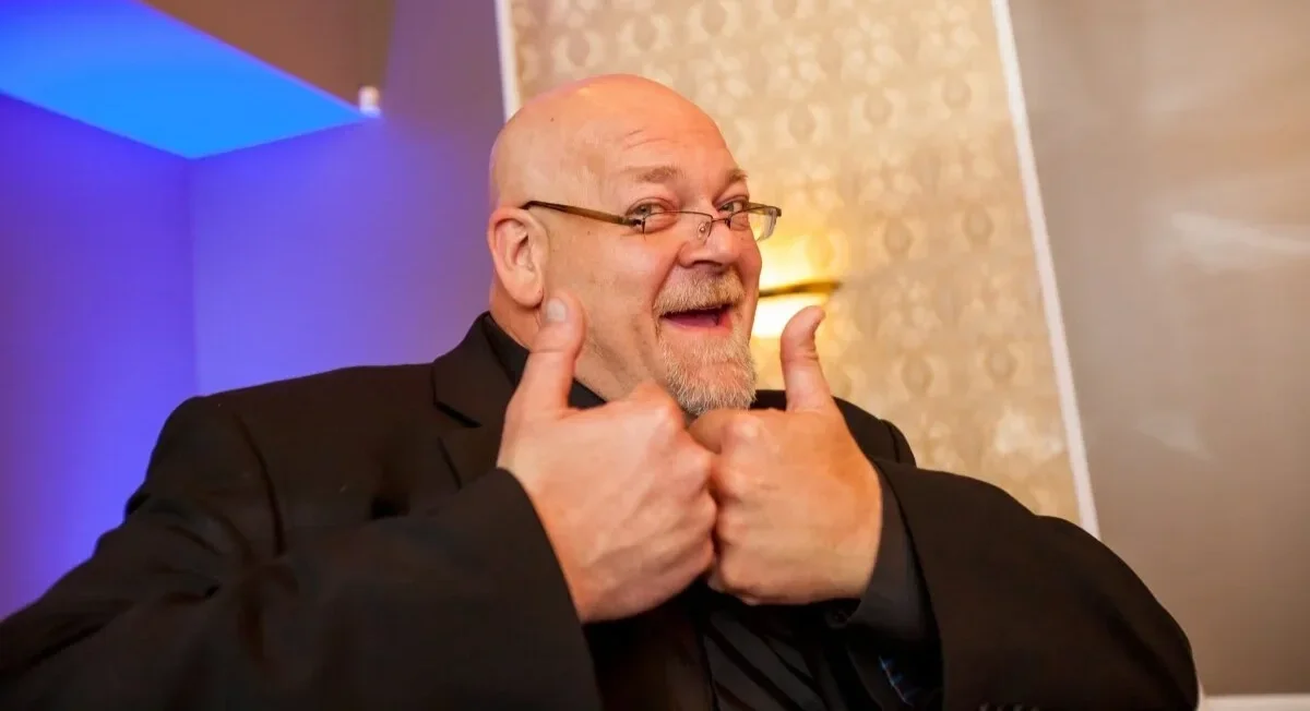 A smiling bald man with glasses and a goatee giving two thumbs up in an indoor setting with colorful lighting.