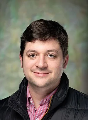 A young man with dark hair, wearing a black jacket over a pink striped shirt, smiling at the camera with a blurred green background.