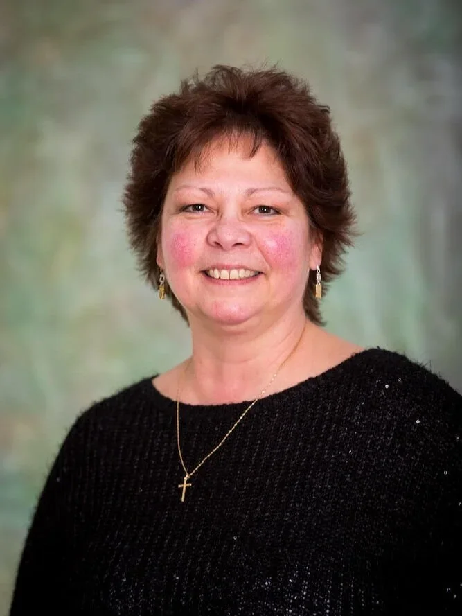 A middle-aged woman with short brown hair, smiling, wearing a black sweater, a gold cross necklace, and gold earrings, standing against a soft green background.