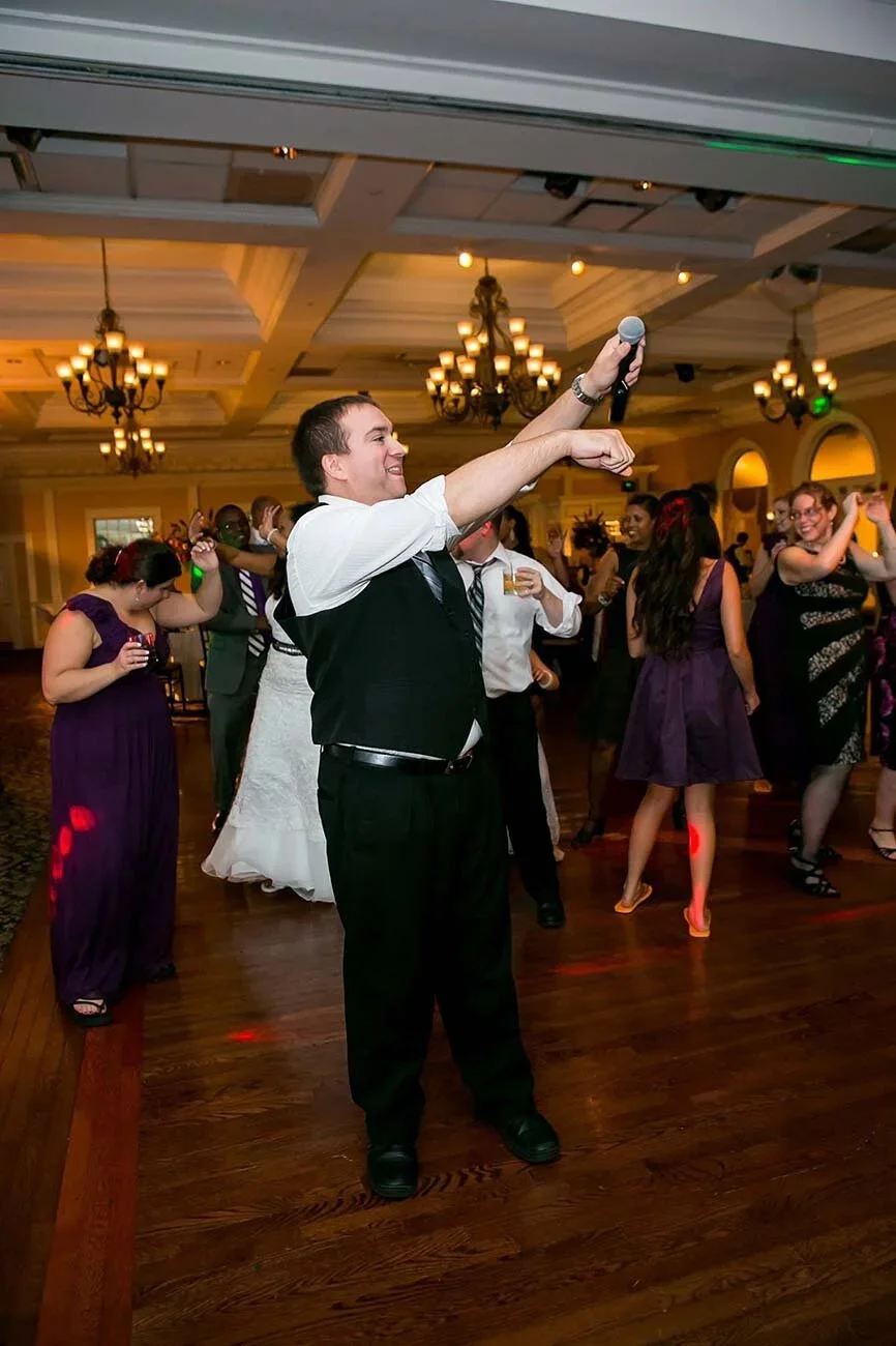 Man dancing and holding a microphone at a party or wedding reception with other guests dancing in the background.