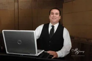 A man in a white shirt and black vest sitting at a desk with a laptop in front of him