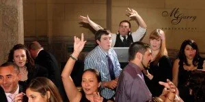 Group of people sitting at a table during a celebration or event, some raising their hands. The background shows a wall with a decorative or scripted sign.