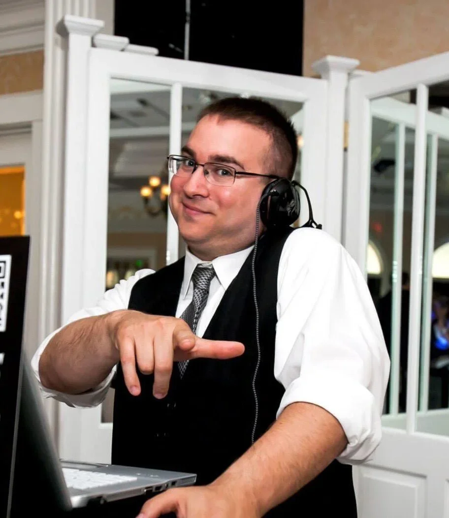 A man wearing glasses and a black vest over a white shirt, with a tie, is pointing at a laptop while wearing a headset. He is smiling and standing in front of a white partition in an indoor setting.
